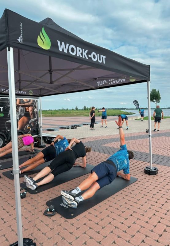 Deelnemers doen core-oefeningen op matten onder een Work-Out tent, met uitzicht op water en natuur.
